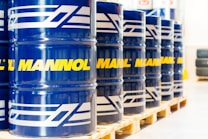 A row of large blue barrels is neatly aligned on wooden pallets. Each barrel prominently features the word 'MANNOL' in bright yellow, with additional white graphics. The scene suggests an industrial or warehouse setting with stacked tires visible in the background.