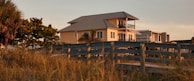 A two-story house with a metal roof and a balcony is set against a backdrop of tropical trees and tall grasses. The lighting suggests sunset, casting a warm glow over the scene. A wooden walkway leads up to the house, enhancing the rustic, coastal setting.
