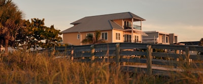 A two-story house with a metal roof and a balcony is set against a backdrop of tropical trees and tall grasses. The lighting suggests sunset, casting a warm glow over the scene. A wooden walkway leads up to the house, enhancing the rustic, coastal setting.
