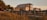 A two-story house with a metal roof and a balcony is set against a backdrop of tropical trees and tall grasses. The lighting suggests sunset, casting a warm glow over the scene. A wooden walkway leads up to the house, enhancing the rustic, coastal setting.