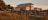 A two-story house with a metal roof and a balcony is set against a backdrop of tropical trees and tall grasses. The lighting suggests sunset, casting a warm glow over the scene. A wooden walkway leads up to the house, enhancing the rustic, coastal setting.