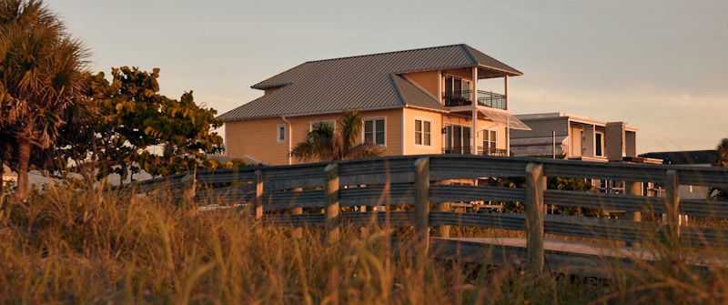 A two-story house with a metal roof and a balcony is set against a backdrop of tropical trees and tall grasses. The lighting suggests sunset, casting a warm glow over the scene. A wooden walkway leads up to the house, enhancing the rustic, coastal setting.