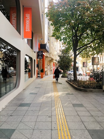 A sidewalk lined with modern buildings featuring large windows and an orange 'OPENHAUS' sign. A person wearing dark clothing walks along the path, carrying an orange bag. A large tree provides shade, and cars are parked on the street.