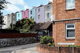 A row of colorful terraced houses with varying shades of gray, blue, pink, and lavender. In the foreground, there is a brick building with a window and a street sign reading 'Lower Cheltenham Place'. There are some bushes and graffiti visible on a wall.