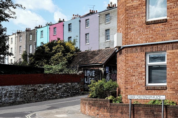 A row of colorful terraced houses with varying shades of gray, blue, pink, and lavender. In the foreground, there is a brick building with a window and a street sign reading 'Lower Cheltenham Place'. There are some bushes and graffiti visible on a wall.