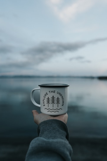 A hand extends holding a metal cup with a motivational phrase 'Explore the Unknown' printed above a graphic of trees and waves. The background shows a calm lake with a serene, cloudy sky blending into a soft horizon.