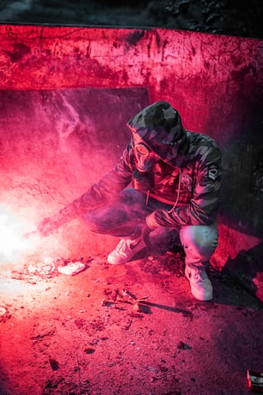A person wearing a hooded camouflage jacket and gas mask crouches on a concrete surface. The scene is illuminated by a reddish flare or smoke, creating a mysterious and intense atmosphere.