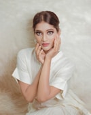 A person with styled hair poses with delicate hands framing their face. The background is a soft, textured fabric, and the individual is wearing a light-colored, elegant outfit.