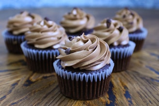 Six chocolate cupcakes topped with swirled chocolate frosting and chocolate chips are arranged on a wooden surface. The cupcakes are in brown liners with a textured appearance. The background is softly blurred, highlighting the focus on the cupcakes.