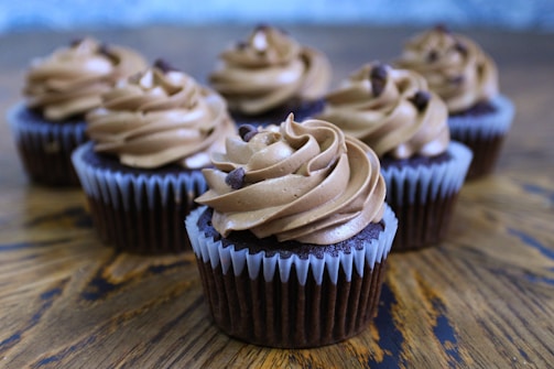 Six chocolate cupcakes topped with swirled chocolate frosting and chocolate chips are arranged on a wooden surface. The cupcakes are in brown liners with a textured appearance. The background is softly blurred, highlighting the focus on the cupcakes.