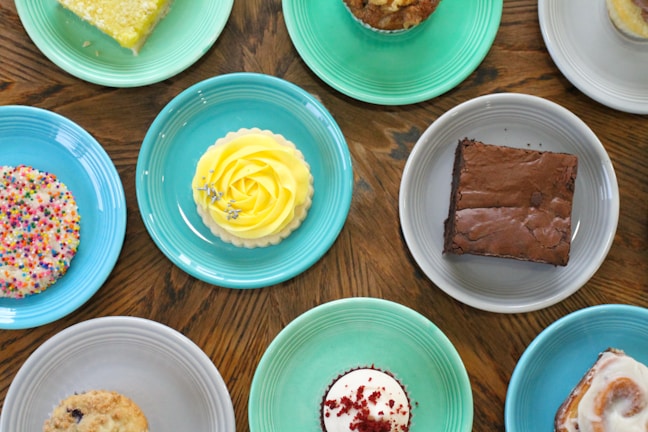 An assortment of desserts is arranged on colorful plates placed on a wooden table. The desserts include a lemon tart with a decorative yellow frosting, a chocolate brownie, a cupcake with red velvet crumbs and white frosting, a large cookie with colorful sprinkles, a muffin, and a cinnamon roll with white icing.