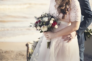 A bride holding a bouquet of flowers, featuring red and white roses, with her groom embracing her from behind. They stand on a beach, indicated by the sand and blurred water in the background. The bride is wearing a lace dress, and the groom is in a dark suit.