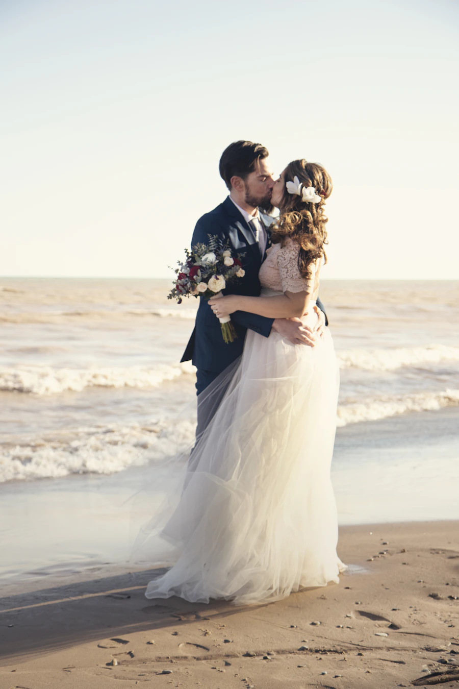 A bride and groom standing together in formal attire beside a calm shoreline