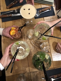 Multiple hands are holding an assortment of cocktails above a wooden table. The drinks include a creamy cocktail with coffee beans on top, two clear drinks with lime slices and mint, and a reddish-brown drink with orange slices. The setting includes cutlery, napkins, and additional drinkware.