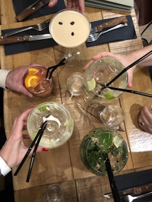Multiple hands are holding an assortment of cocktails above a wooden table. The drinks include a creamy cocktail with coffee beans on top, two clear drinks with lime slices and mint, and a reddish-brown drink with orange slices. The setting includes cutlery, napkins, and additional drinkware.