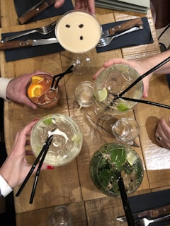 Multiple hands are holding an assortment of cocktails above a wooden table. The drinks include a creamy cocktail with coffee beans on top, two clear drinks with lime slices and mint, and a reddish-brown drink with orange slices. The setting includes cutlery, napkins, and additional drinkware.