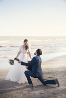 A romantic scene at the beach with a couple engaged in what appears to be a proposal. The woman is dressed in a flowing white wedding gown, holding a bouquet of flowers, and the man is kneeling in a navy blue suit, holding a guitar. The background features a calm ocean and a clear sky.