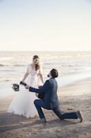 A romantic scene at the beach with a couple engaged in what appears to be a proposal. The woman is dressed in a flowing white wedding gown, holding a bouquet of flowers, and the man is kneeling in a navy blue suit, holding a guitar. The background features a calm ocean and a clear sky.