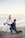 A romantic scene at the beach with a couple engaged in what appears to be a proposal. The woman is dressed in a flowing white wedding gown, holding a bouquet of flowers, and the man is kneeling in a navy blue suit, holding a guitar. The background features a calm ocean and a clear sky.