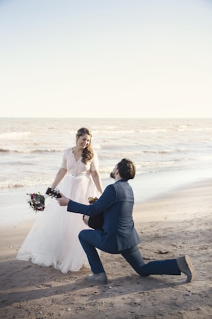 A romantic scene at the beach with a couple engaged in what appears to be a proposal. The woman is dressed in a flowing white wedding gown, holding a bouquet of flowers, and the man is kneeling in a navy blue suit, holding a guitar. The background features a calm ocean and a clear sky.