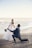 A romantic scene at the beach with a couple engaged in what appears to be a proposal. The woman is dressed in a flowing white wedding gown, holding a bouquet of flowers, and the man is kneeling in a navy blue suit, holding a guitar. The background features a calm ocean and a clear sky.