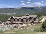A large wooden lodge set against a backdrop of lush green forests and distant mountains. The building has multiple stories with dormer windows and a wide, steeply sloped roof. The area around the lodge is lively, with visible people and outdoor activities scattered across a grassy hillside. Paved pathways and recreational areas are present, suggesting a resort or vacation setting.