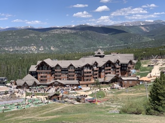 A large wooden lodge set against a backdrop of lush green forests and distant mountains. The building has multiple stories with dormer windows and a wide, steeply sloped roof. The area around the lodge is lively, with visible people and outdoor activities scattered across a grassy hillside. Paved pathways and recreational areas are present, suggesting a resort or vacation setting.