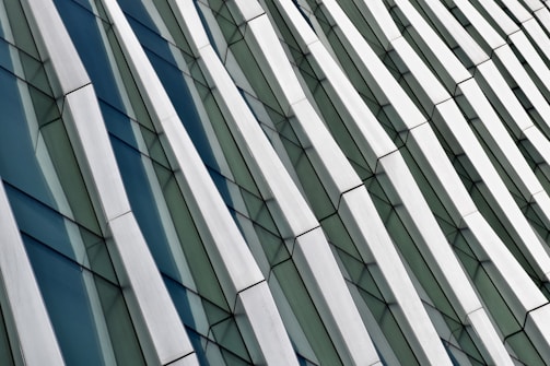The image depicts an abstract architectural facade featuring a series of angular, overlapping panels with a reflective quality. The repetitive pattern creates a sense of movement and depth with a combination of glass and metallic surfaces.