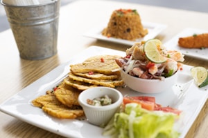 A plate with fried plantains, a bowl of seafood salad garnished with lime, and sides of lettuce and sliced tomatoes. In the background, there is a serving of rice with vegetables and a piece of fried food on separate plates. A metal bucket, possibly for utensils, is partially visible.