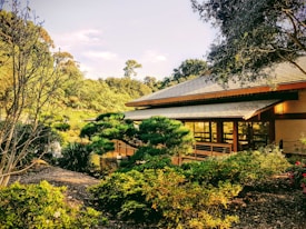 A serene garden setting with a traditional Japanese-style building, surrounded by lush greenery and manicured bushes. The architecture features a sloping roof with wooden beams, and large windows reflecting the surrounding landscape. Sunlight filters through the trees, casting a warm glow over the scene.