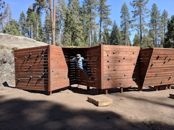 A person is climbing on a wooden bouldering wall outdoors, surrounded by tall pine trees. The structure is made of wooden panels with climbing holds attached. Sunlight creates shadows on the ground, and the area is covered with dirt.