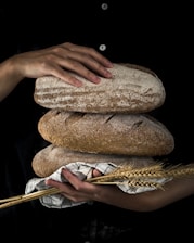 A person is holding three loaves of rustic bread, one resting on top of the others. A hand rests gently on the top loaf. The loaves feature a crusty texture and appear artisanal. A few stalks of wheat and a striped cloth are included in the arrangement, suggesting a connection to traditional baking or agriculture.