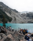 A group of people gathered on rocky terrain by a stunning turquoise alpine lake, with a backdrop of rugged mountains. The atmosphere is lively with individuals exploring and taking in the scenic landscape. A pine tree stands on the left, and a glacier is visible in the distance.