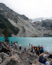 A group of people gathered on rocky terrain by a stunning turquoise alpine lake, with a backdrop of rugged mountains. The atmosphere is lively with individuals exploring and taking in the scenic landscape. A pine tree stands on the left, and a glacier is visible in the distance.