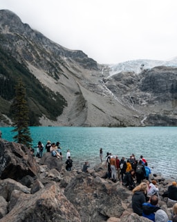A group of people gathered on rocky terrain by a stunning turquoise alpine lake, with a backdrop of rugged mountains. The atmosphere is lively with individuals exploring and taking in the scenic landscape. A pine tree stands on the left, and a glacier is visible in the distance.