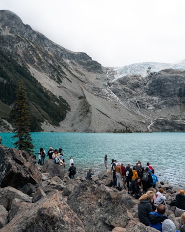 A group of people gathered on rocky terrain by a stunning turquoise alpine lake, with a backdrop of rugged mountains. The atmosphere is lively with individuals exploring and taking in the scenic landscape. A pine tree stands on the left, and a glacier is visible in the distance.