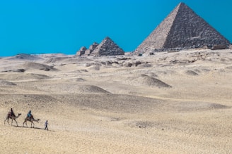 In a vast desert landscape, two large pyramids stand prominently in the background under a clear blue sky. The foreground features undulating sand dunes with two camels being led by riders, accompanied by a person walking beside them.