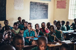 A classroom filled with young students wearing uniforms in shades of green and blue. The walls are adorned with chalkboards and posters, and natural light filters through a window. The students are seated at wooden desks, some are smiling while others look attentive.