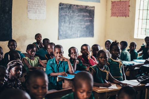 A classroom filled with young students wearing uniforms in shades of green and blue. The walls are adorned with chalkboards and posters, and natural light filters through a window. The students are seated at wooden desks, some are smiling while others look attentive.