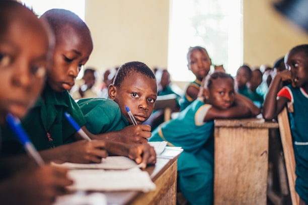 A group of children sitting at wooden desks in a classroom environment. They are focused on writing with pens, and wearing green uniforms. The room appears well-lit with natural light coming through a window.