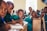 A group of children sitting at wooden desks in a classroom environment. They are focused on writing with pens, and wearing green uniforms. The room appears well-lit with natural light coming through a window.