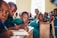 A group of children sitting at wooden desks in a classroom environment. They are focused on writing with pens, and wearing green uniforms. The room appears well-lit with natural light coming through a window.