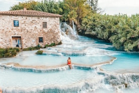 A serene landscape featuring a series of tiered, turquoise-blue natural hot springs with a rustic stone building to the left. The building has multiple windows and a wooden door, surrounded by verdant foliage. A person in a red swimsuit sits at the edge of one of the pools, enjoying the tranquil environment. The cascading water creates a soothing effect, with lush greenery surrounding the scene.