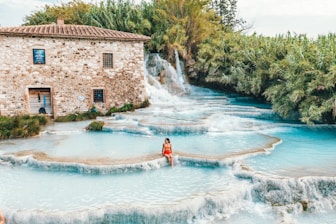 A serene landscape featuring a series of tiered, turquoise-blue natural hot springs with a rustic stone building to the left. The building has multiple windows and a wooden door, surrounded by verdant foliage. A person in a red swimsuit sits at the edge of one of the pools, enjoying the tranquil environment. The cascading water creates a soothing effect, with lush greenery surrounding the scene.