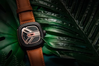 A stylish wristwatch with a brown leather strap rests on top of large, glossy green tropical leaves. The watch face displays intricate details with a blend of silver and black elements, and the name 'Seven Friday' is visible.