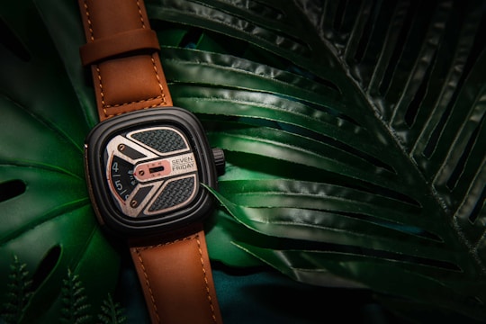 A stylish wristwatch with a brown leather strap rests on top of large, glossy green tropical leaves. The watch face displays intricate details with a blend of silver and black elements, and the name 'Seven Friday' is visible.