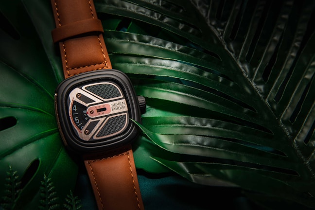A stylish wristwatch with a brown leather strap rests on top of large, glossy green tropical leaves. The watch face displays intricate details with a blend of silver and black elements, and the name 'Seven Friday' is visible.