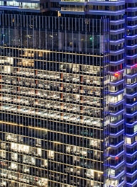 A high-rise office building with a grid of illuminated windows, showing numerous office spaces inside. The structure features modern architectural design with blue accent lighting along the edges and a mix of warm interior lights.