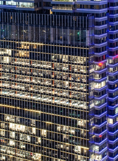 A high-rise office building with a grid of illuminated windows, showing numerous office spaces inside. The structure features modern architectural design with blue accent lighting along the edges and a mix of warm interior lights.