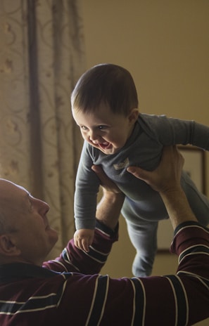 man wearing maroon, white, and blue stripe long-sleeved shirt lifting up baby wearing gray onesie